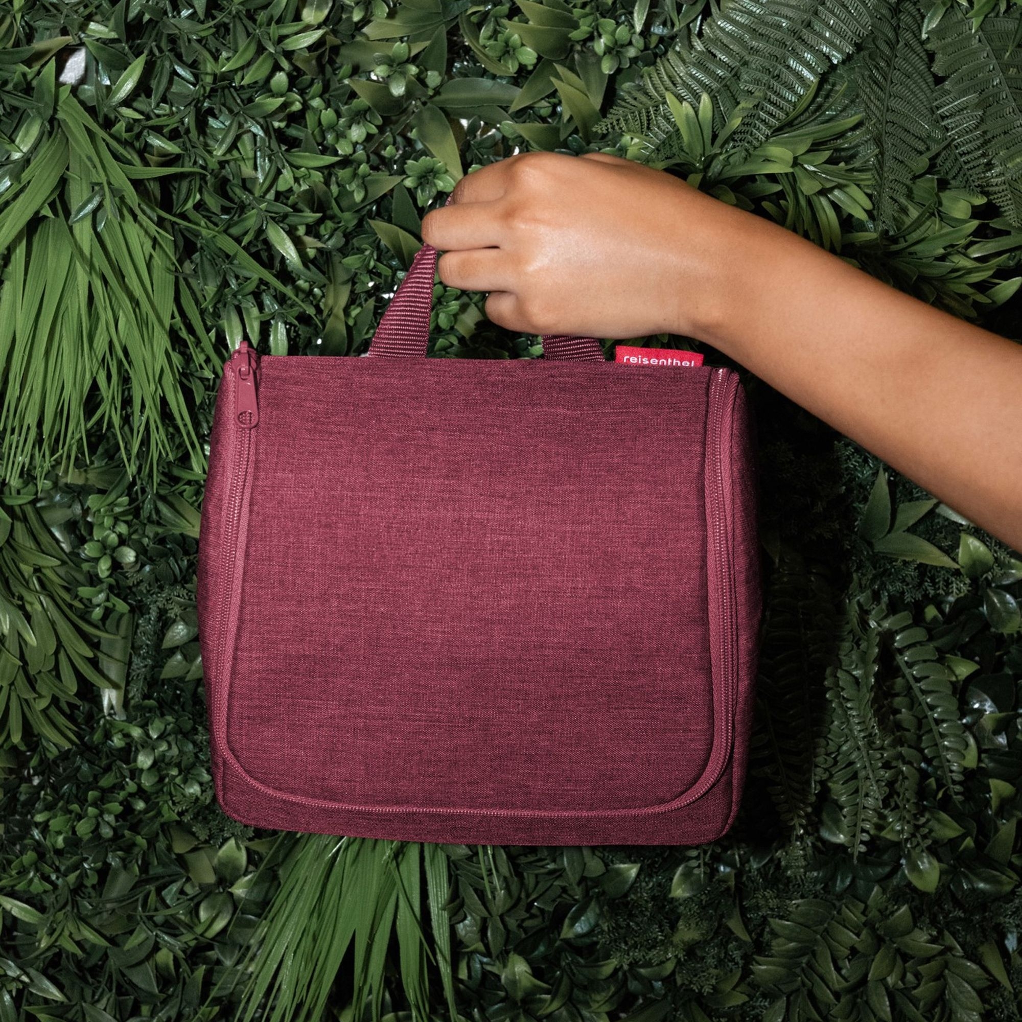 Hand holding burgundy travel bag in front of green plants