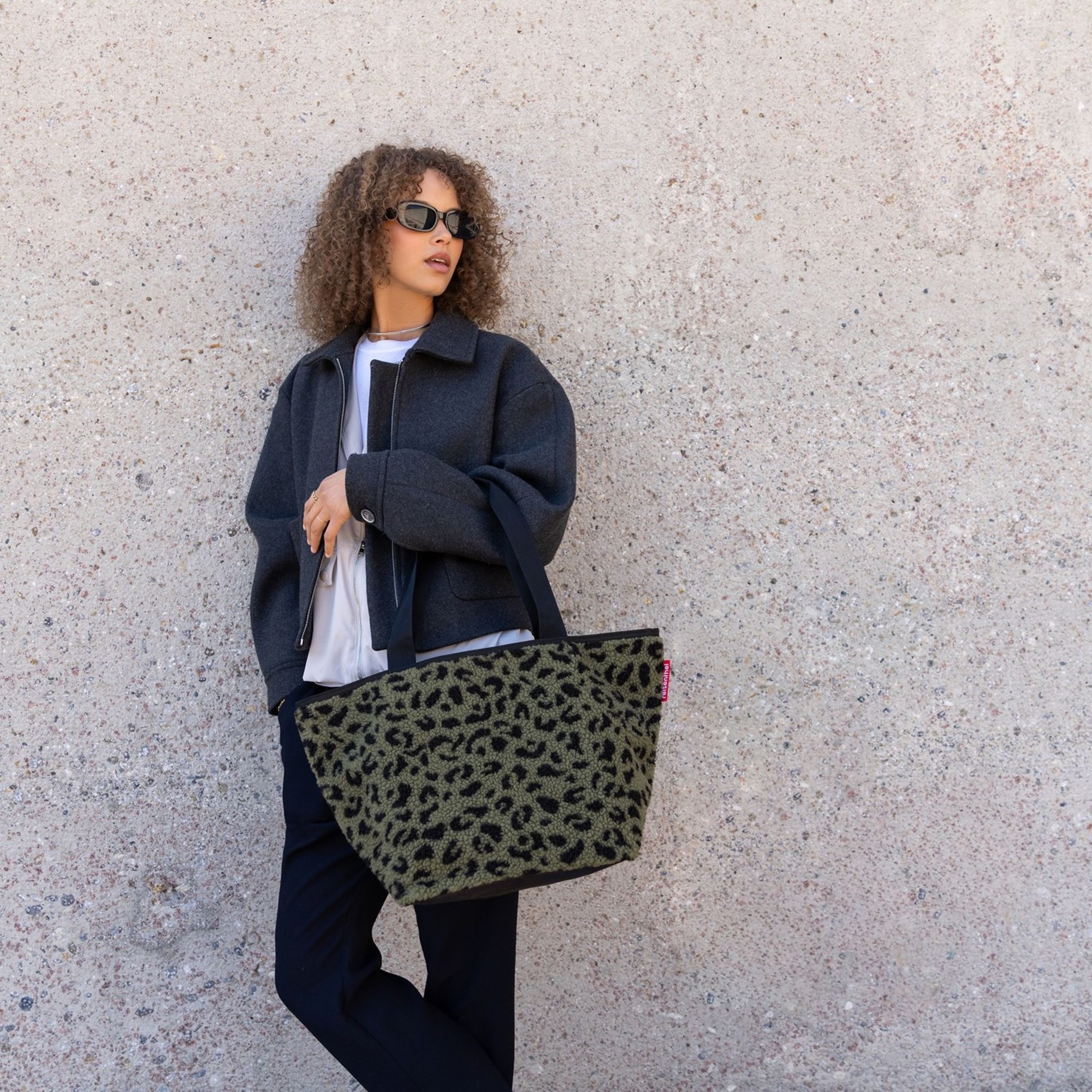 Woman with dark jacket and large leopard bag by wall